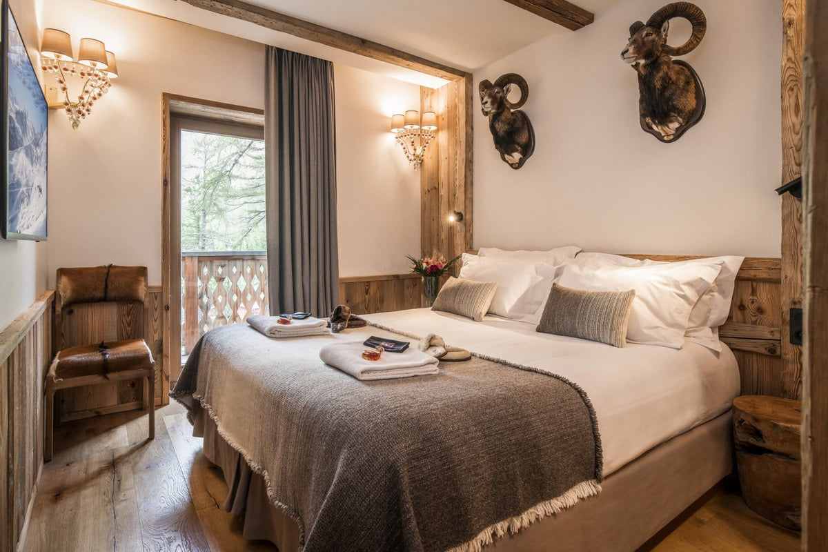 A rustic bedroom interior with a large bed dressed in white linens and a gray throw blanket, set in a cabin-like room with wood walls, exposed wooden beams, wall-mounted sconces, a chair by a window, and two mounted ram heads on the wall.