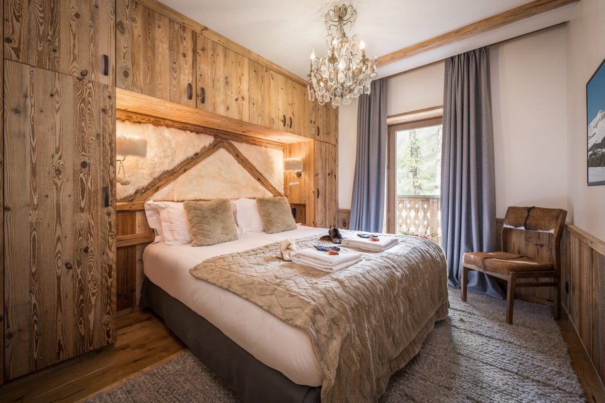 A rustic alpine-style bedroom featuring a large bed dressed in white and beige linens, set against a wall of natural wood cabinetry. A crystal chandelier hangs from the ceiling, and a window with gray curtains looks out to a balcony.