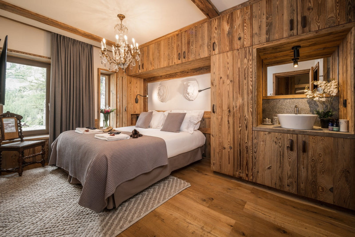 A rustic bedroom interior with a large bed dressed in white and taupe bedding, surrounded by wood-paneled walls, a chandelier, and a built-in vanity area with a sink and mirror.