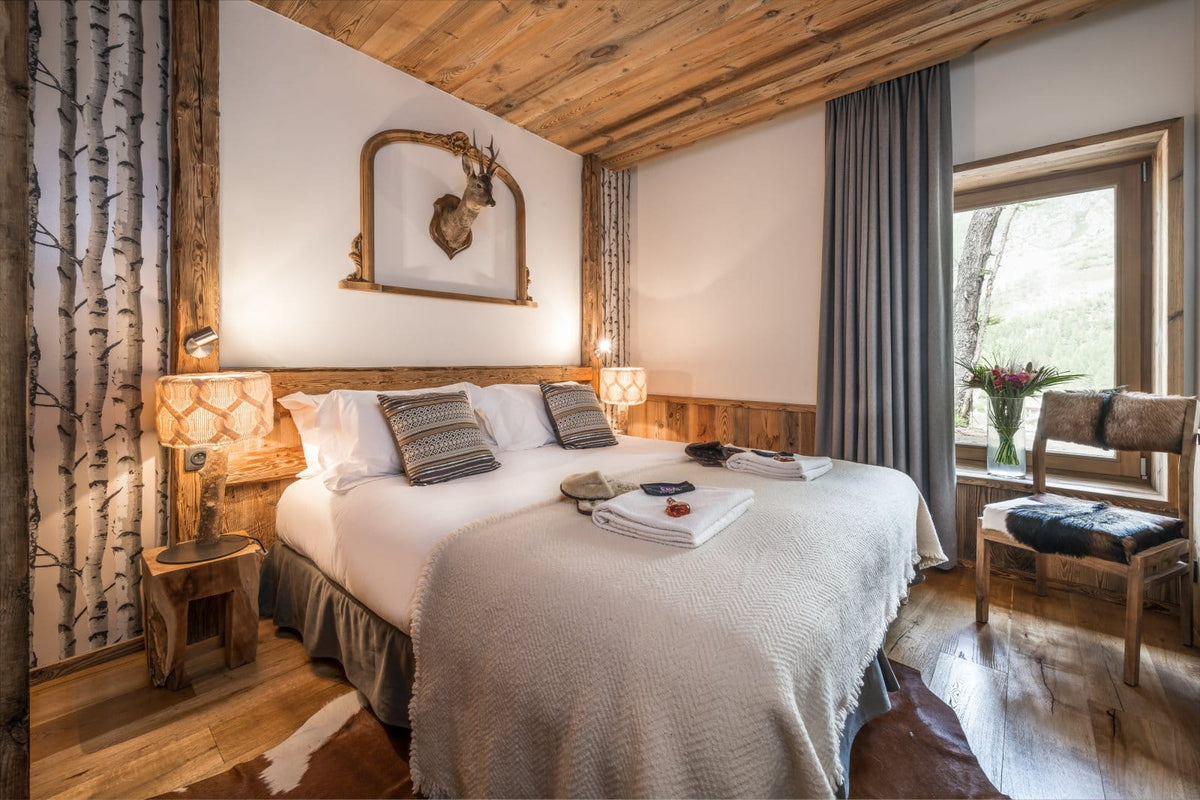 A rustic bedroom interior with a neatly made bed covered in white bedding and textured beige throws, set in a wood-paneled room with bedside lamps, a window, curtains, and a chair.