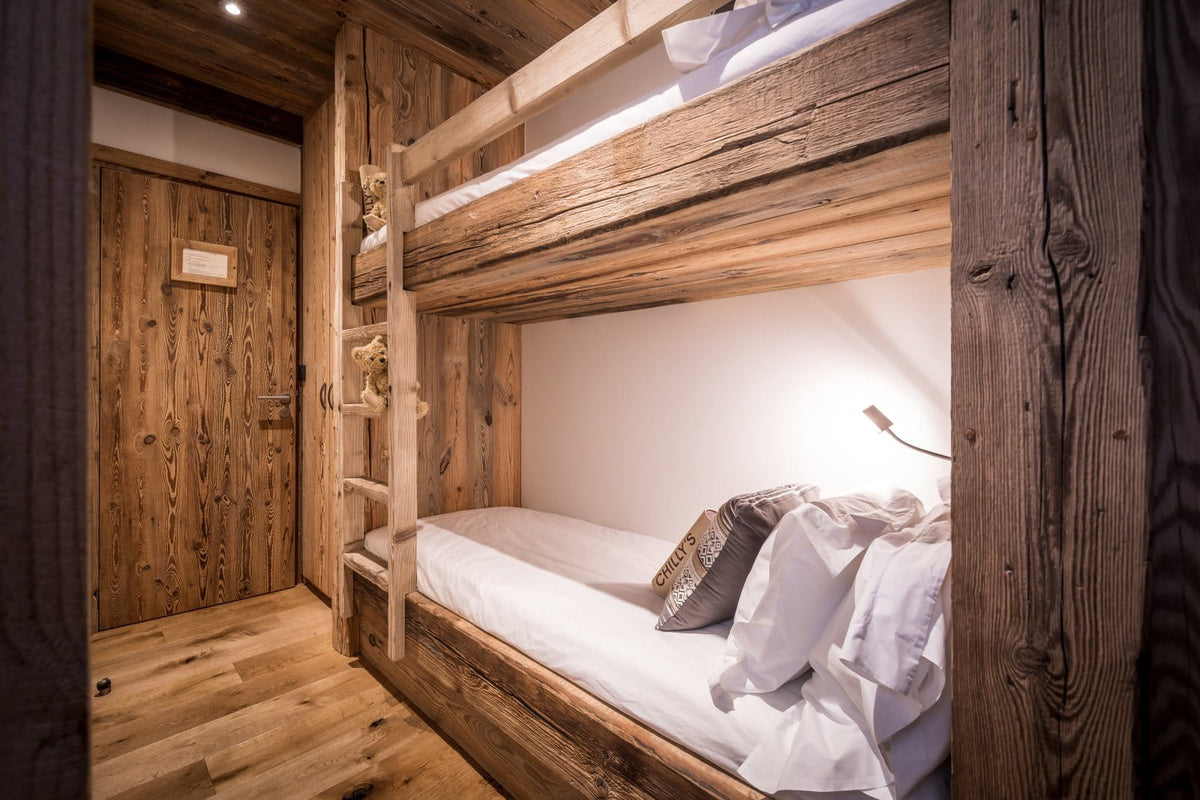 A rustic wooden bunk bed with white bedding in a cozy, wood-paneled bedroom. A small wall-mounted reading light illuminates the lower bed, and a matching wood door is visible in the background.