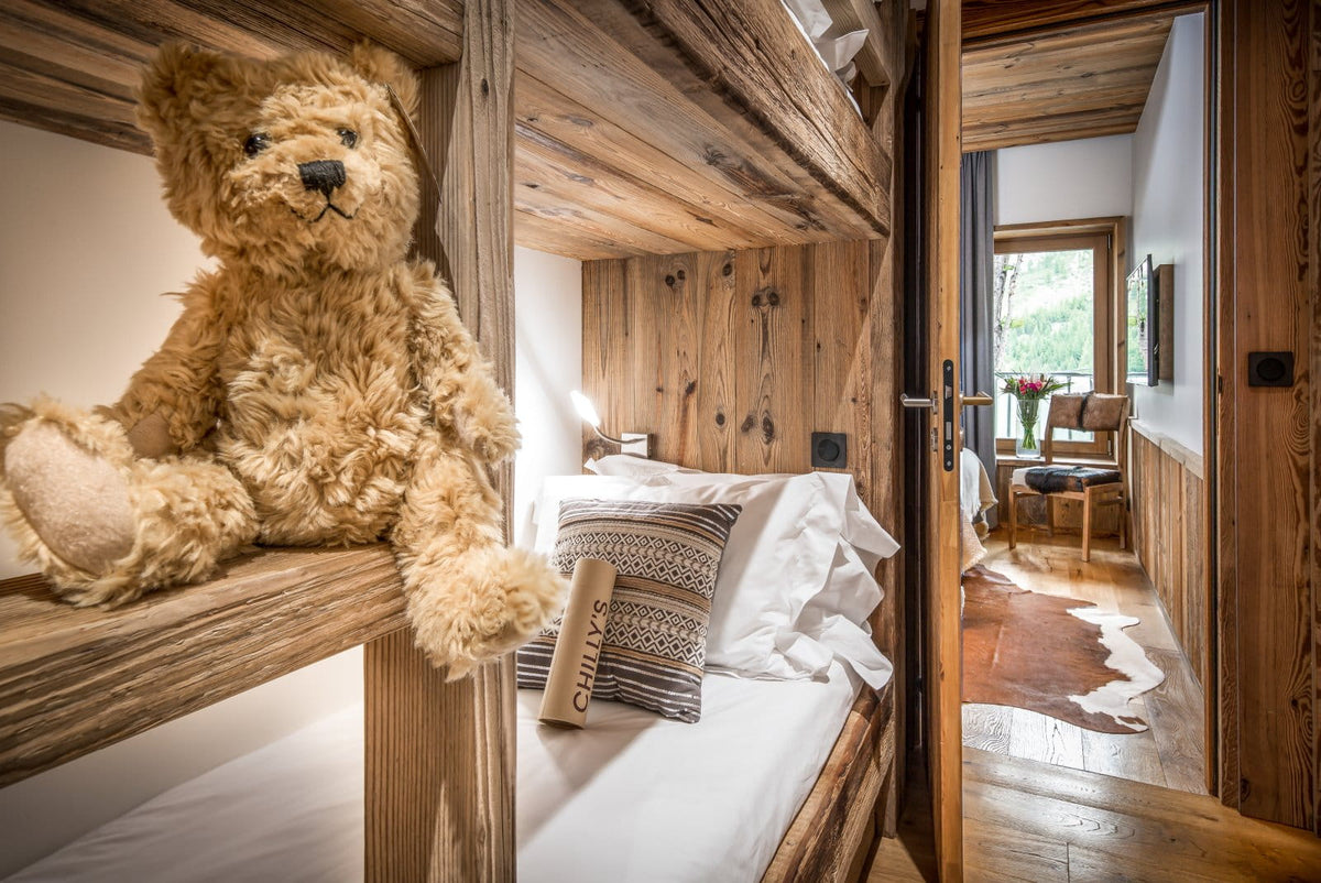 A large fluffy teddy bear sits on a rustic wooden bunk bed frame in a cozy alpine-style bedroom with wood paneling, white bedding, and an open doorway leading to another room.