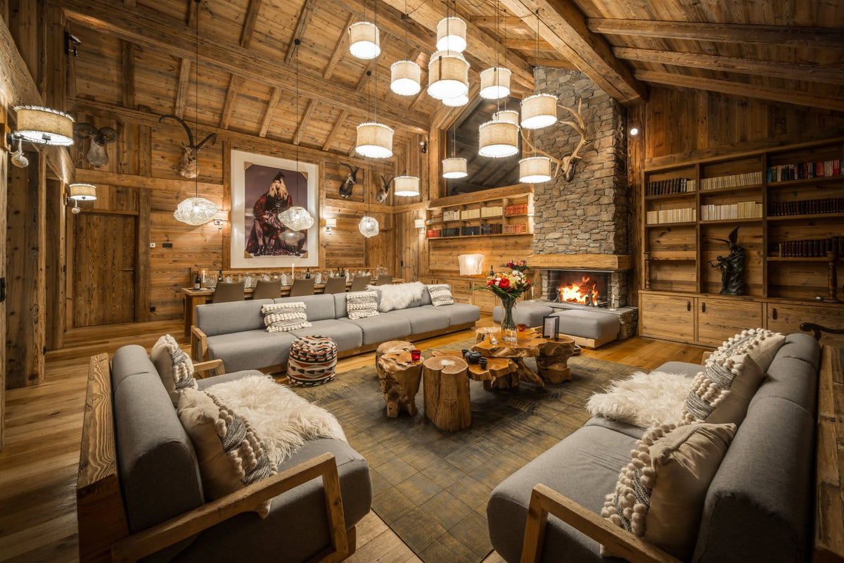 A cozy, rustic chalet-style living room with gray sofas, a stone fireplace, wooden ceiling beams, and multiple hanging pendant lights.