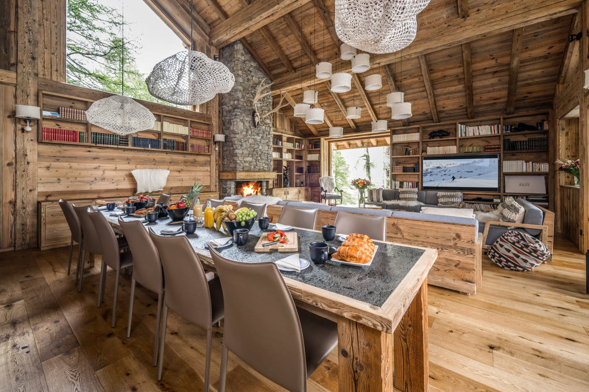 A rustic alpine chalet interior with a long dining table set for guests, upholstered chairs, pendant lights, wood-paneled walls, a stone fireplace, and a living area in the background.