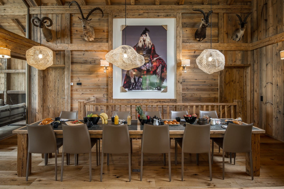 A rustic wooden dining room with a long wooden table, ten gray upholstered chairs, three woven pendant lights, mounted animal heads and horns on the wood-paneled walls, and a large framed portrait hanging in the center.