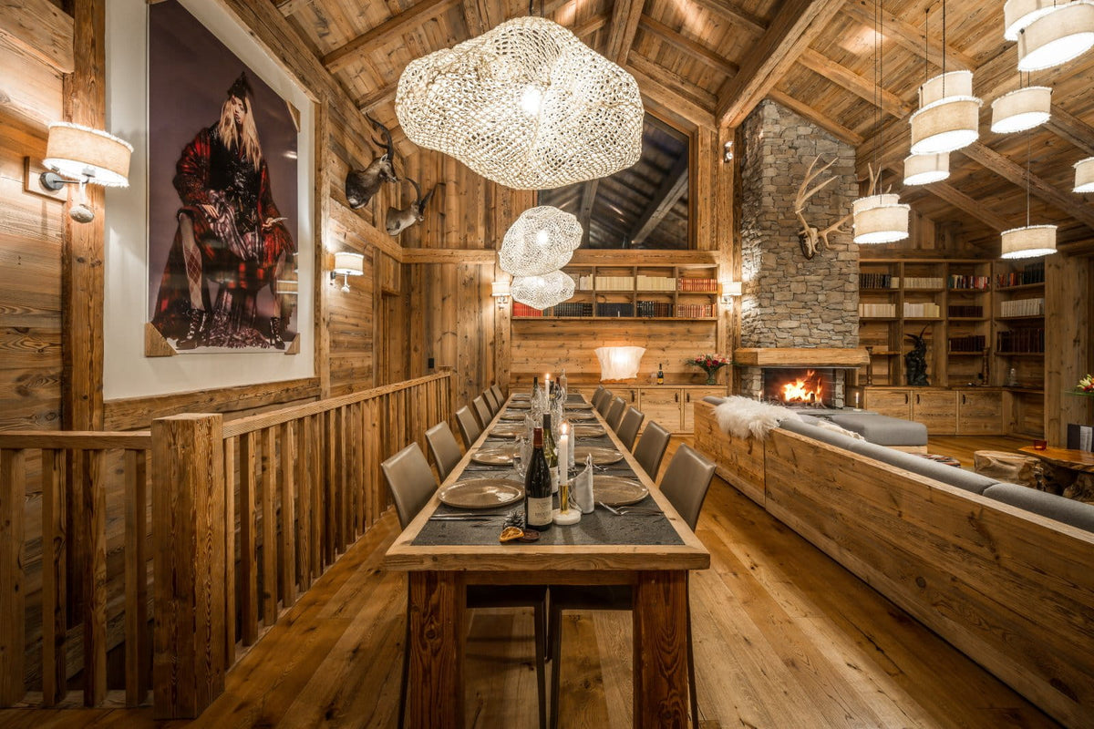 A rustic alpine chalet dining room with a long wooden dining table set for six, surrounded by matching chairs. The room features a high wood-beamed ceiling, warm ambient lighting from multiple hanging pendant lamps, a stone fireplace with a fire burning in the background, and a large framed portrait on the left wall.