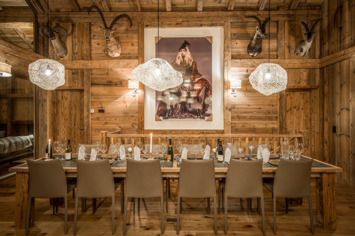 A rustic wooden dining room with a long table set for multiple guests, hanging woven pendant lights, mounted animal heads on the wood-paneled walls, and a large framed artwork centered above the table.