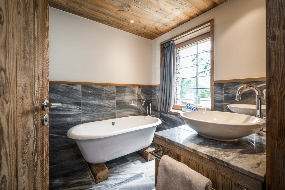A rustic alpine-style bathroom with a freestanding white bathtub on wooden blocks, a marble countertop with a white vessel sink, dark gray marble wall tiles, and a wooden ceiling.
