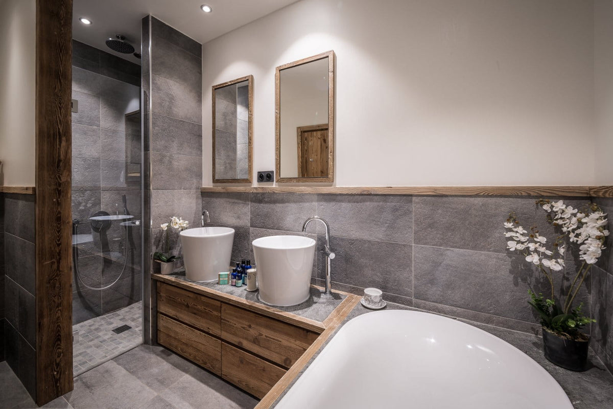A modern bathroom interior with a double white vessel sinks on a wooden vanity, a white freestanding bathtub in the foreground, gray stone tile walls and floor, two wood-framed mirrors, and a glass shower enclosure.