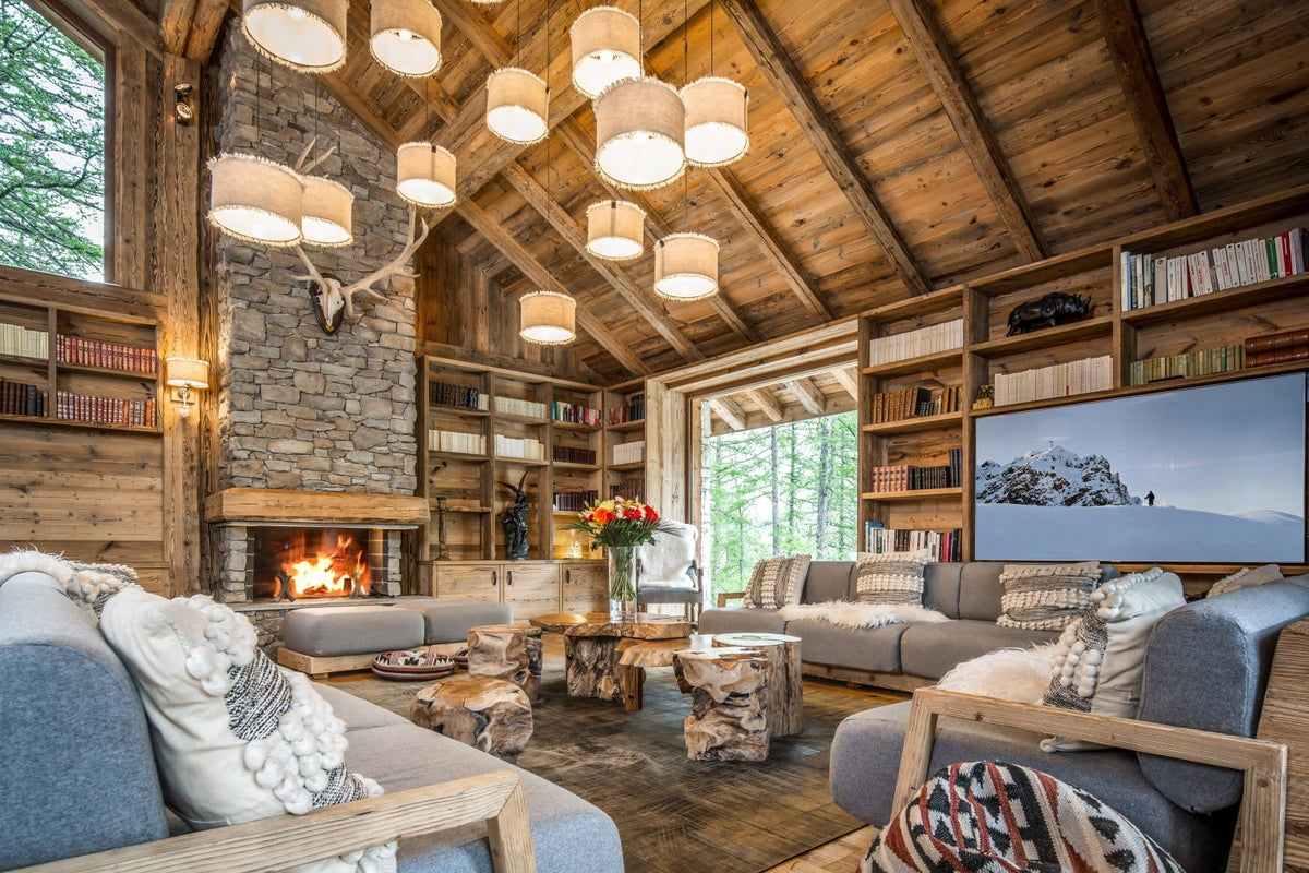 A cozy ski chalet living room with a high wood-beamed ceiling, stone fireplace, built-in bookshelves, gray sofas, rustic log tables, and multiple hanging drum pendant lights. A large TV on the right wall displays a snowy mountain scene.