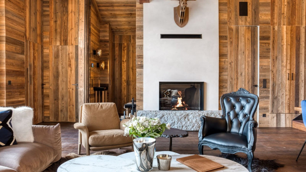 A rustic, cozy living room with extensive warm wood-paneled walls, featuring a fireplace, a black leather tufted armchair, a beige armchair, a sofa, and a round marble coffee table in the foreground.