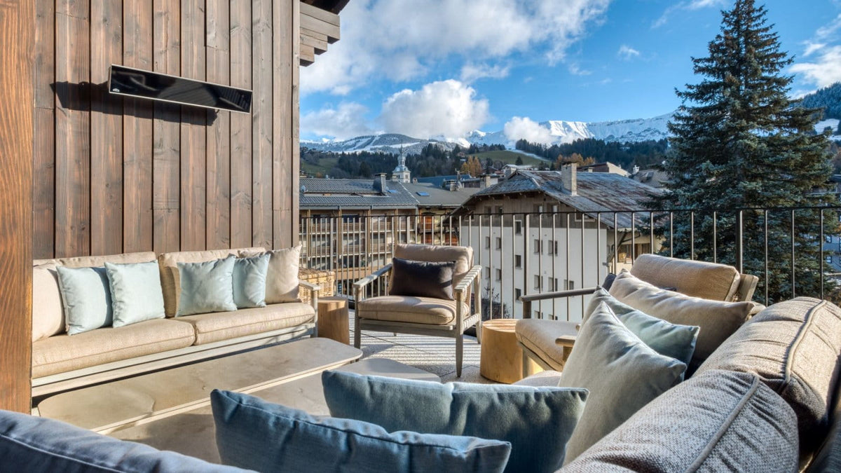 Outdoor patio seating area with beige sofas, blue accent pillows, a wooden wall, and a balcony railing overlooking a mountain village under a blue sky.
