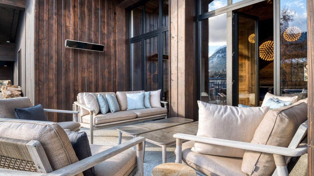 Modern outdoor patio furniture set with beige and light blue cushions, woven wicker armchairs, and a low wooden coffee table, staged on a deck with dark wood paneling and large glass doors.