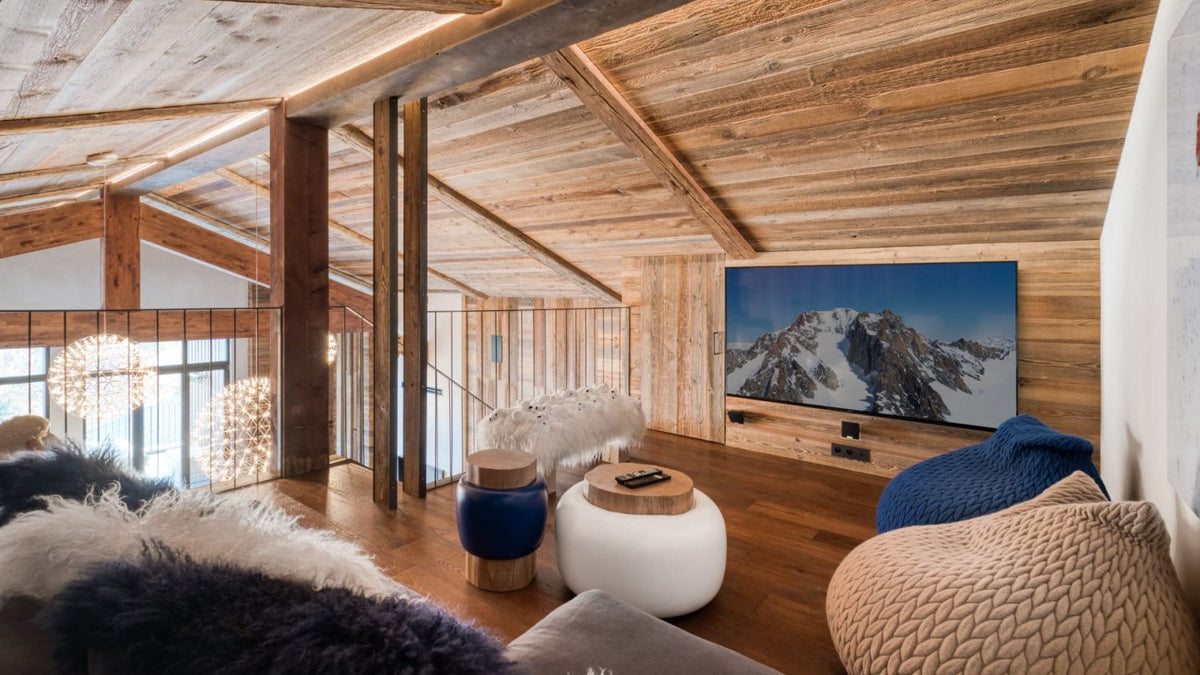 A modern rustic mountain chalet living room with a large wall-mounted TV displaying a snowy mountain landscape. The room features a warm wood ceiling, wooden wall panels, a wooden floor, a white round coffee table, a blue side table, and cozy textured bean bags in the foreground.