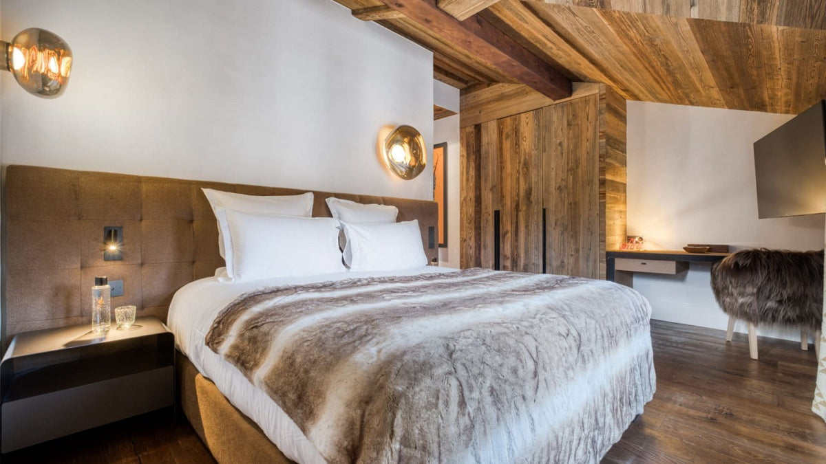 A luxury bedroom featuring a large bed with white linens and a brown faux-fur throw, set against a white wall with a brown leather headboard. The room has rustic wooden ceiling beams, a matching wooden wardrobe, and a desk area with a chair.
