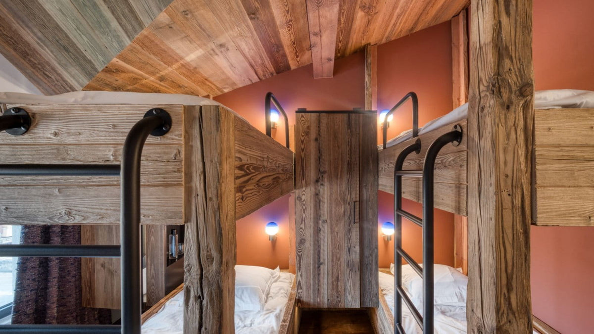 A rustic wooden bunk bed frame with black metal ladder rails in a slanted-roof bedroom, with peach-colored walls and warm ambient lighting.