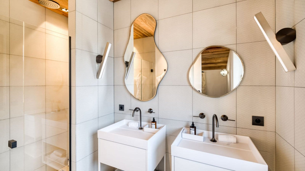 Modern bathroom interior with two white sinks, irregular and round mirrors, black faucets, wall-mounted light fixtures, beige tiled walls, and a glass shower enclosure.