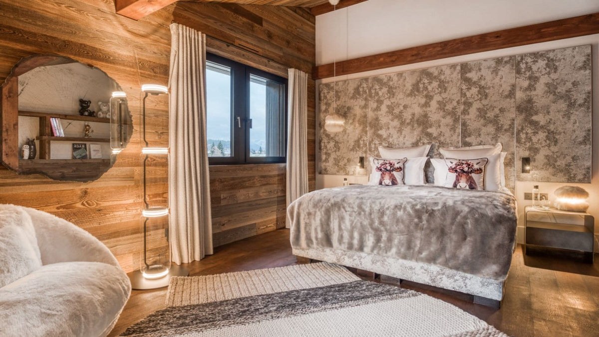 A luxury bedroom interior with a large bed dressed in gray and white textured bedding, set against a patterned accent wall. The room features rustic wood-paneled walls, a large rustic wall mirror, a modern floor lamp, and a cozy rug.