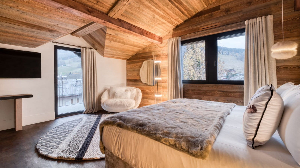 A cozy mountain chalet bedroom featuring a large bed with a textured gray fur throw, white bedding, and a round patterned rug. The room has warm wood-paneled walls and ceiling with exposed wooden beams, large windows with white curtains, and a modern white armchair.