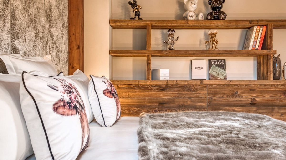 A cozy bedroom scene featuring a bed with white pillows, some with a printed animal design, a gray faux-fur throw blanket, and a rustic wooden headboard and shelving unit in the background.
