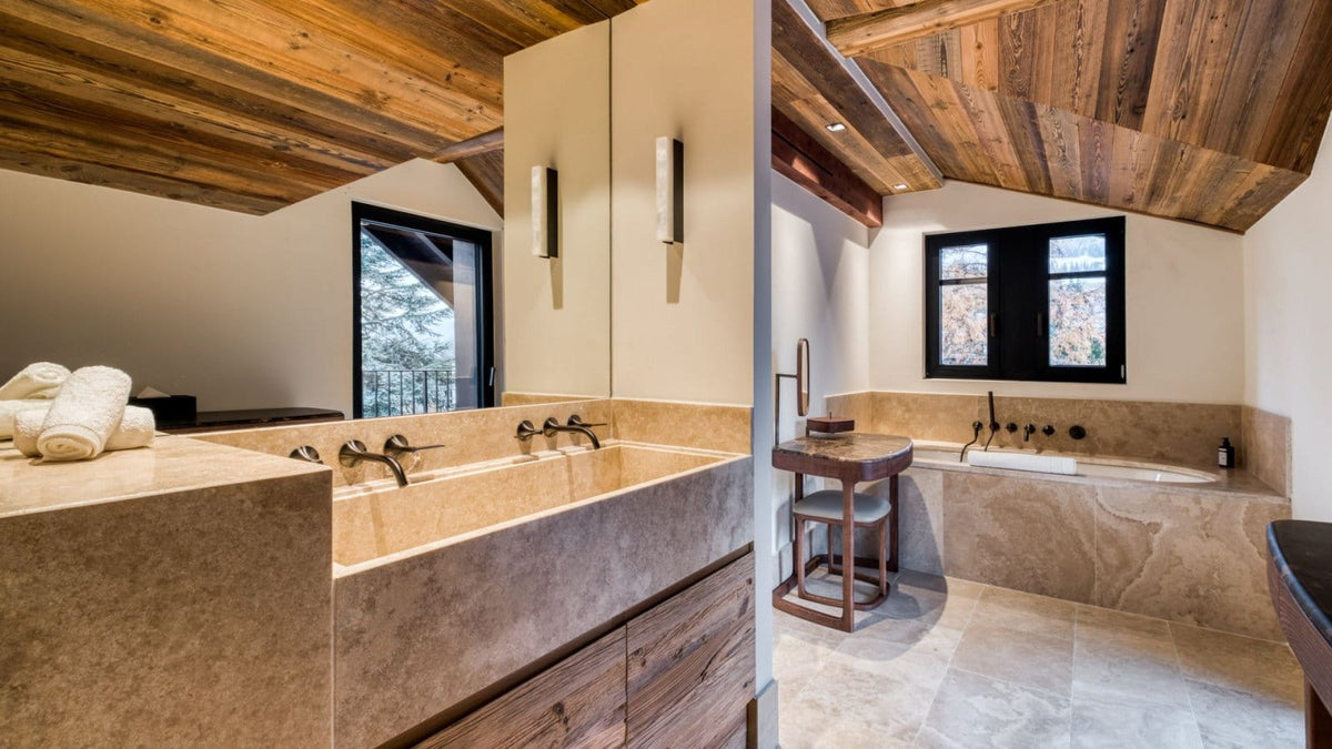 A luxury spa-style bathroom interior featuring a large stone double vanity with a mirror, a built-in bathtub, a small wooden stool, and a rustic wood-beamed ceiling.