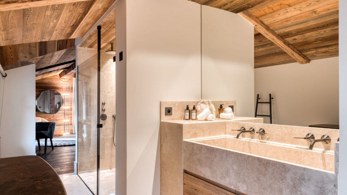 A modern bathroom interior with a stone double vanity, large wall mirror, glass shower enclosure, and warm wood ceiling beams.
