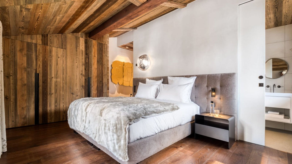 A modern bedroom featuring a bed with white linens and a gray textured throw, set against a white wall with a round wall lamp. The room has a prominent rustic wooden ceiling and matching wood-paneled walls, dark wood flooring, and a sleek black nightstand.