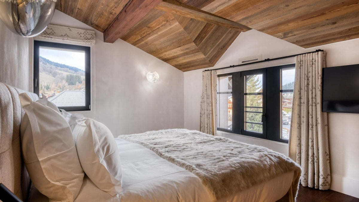A luxury bedroom featuring a large bed with white linens and a textured beige throw, set against a rustic wood-beamed ceiling and white walls. Two windows with black frames offer a view of a mountain landscape, dressed with patterned cream curtains.