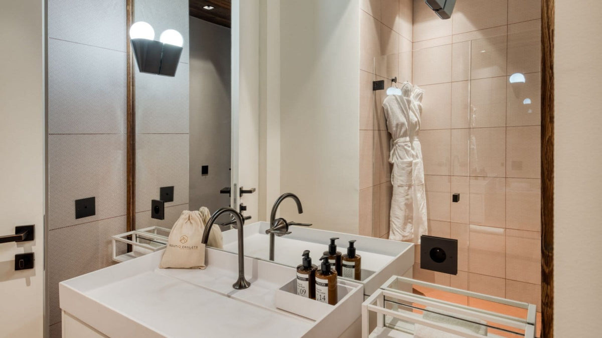 A modern bathroom vanity with a white double sink, matte black faucets, and a large mirror. A white robe hangs on a rack in the tiled shower area behind, and a small beige toiletry bag sits on the counter.