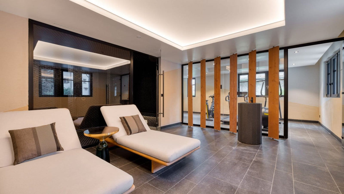 A modern indoor lounge area with two white chaise lounge chairs, gray floor tiles, warm wood partition panels, recessed ceiling lighting, and a glass-walled interior space in the background.
