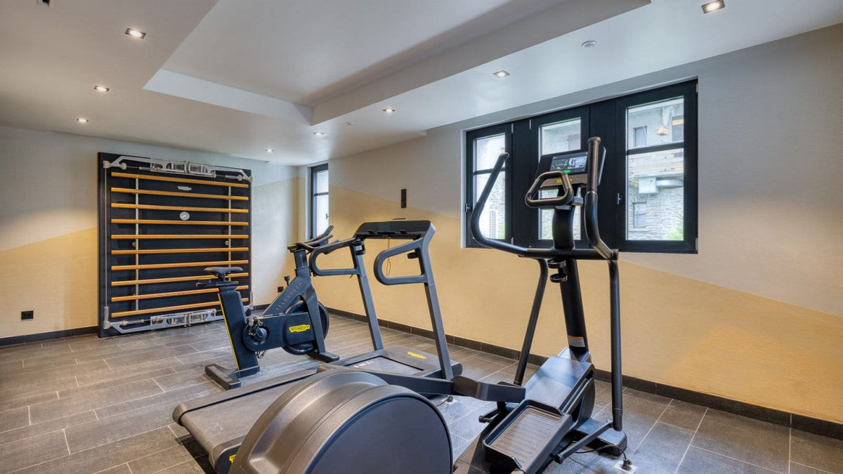 A modern home gym interior featuring a treadmill, an elliptical machine, and a stationary bike, set against beige and yellow walls with dark gray tiled flooring.