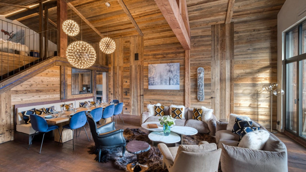 A luxury mountain chalet living room with rustic wood-paneled walls and ceiling, featuring a dining area with blue chairs, a cozy seating area with beige sofas, and spherical woven pendant lights.