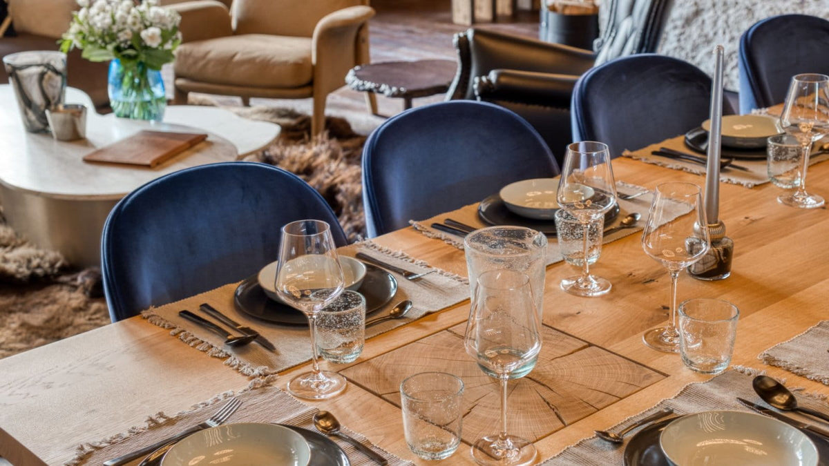 A styled dining table set with clear drinking glasses, plates, cutlery, and placemats on a light wood surface, surrounded by blue upholstered chairs in a warm, furnished interior.