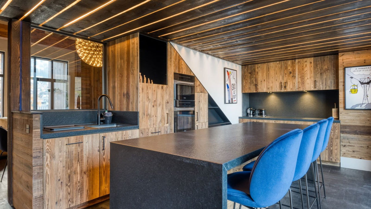 A modern kitchen interior with wood cabinetry, a dark countertop island, blue bar stools, and a slanted ceiling with linear lighting.