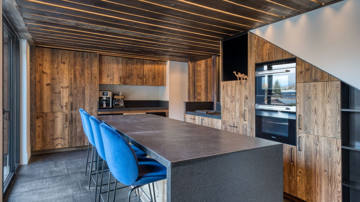 A modern kitchen interior with wood-paneled cabinets, a dark stone island, built-in stainless steel appliances, and three blue bar stools in the foreground.