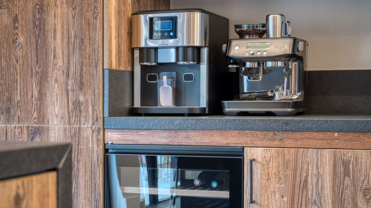 A modern kitchen setup featuring a stainless steel coffee machine and a countertop beverage dispenser on a dark stone surface, set against warm wood grain cabinets.