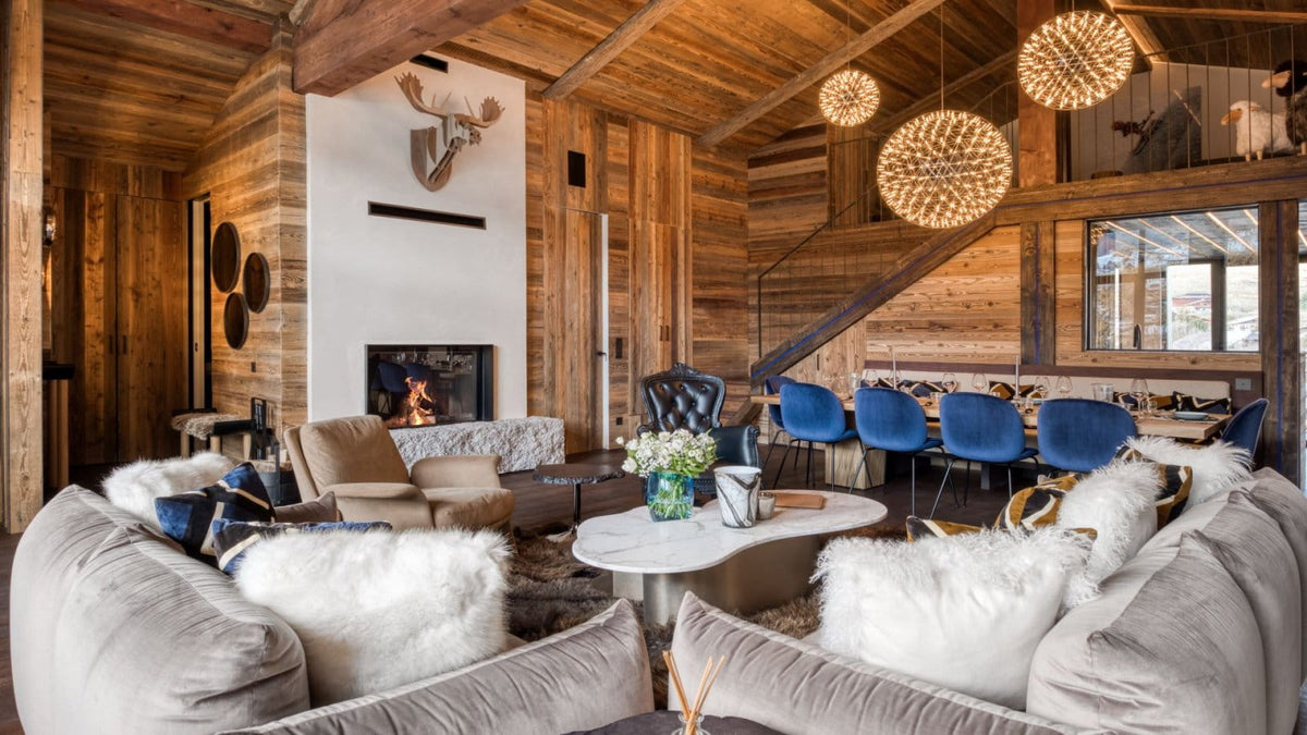 A luxurious ski chalet living room featuring a large gray sectional sofa with white sheepskin pillows, a round coffee table, blue accent chairs, a fireplace, and a wooden staircase under a high ceiling with exposed beams.