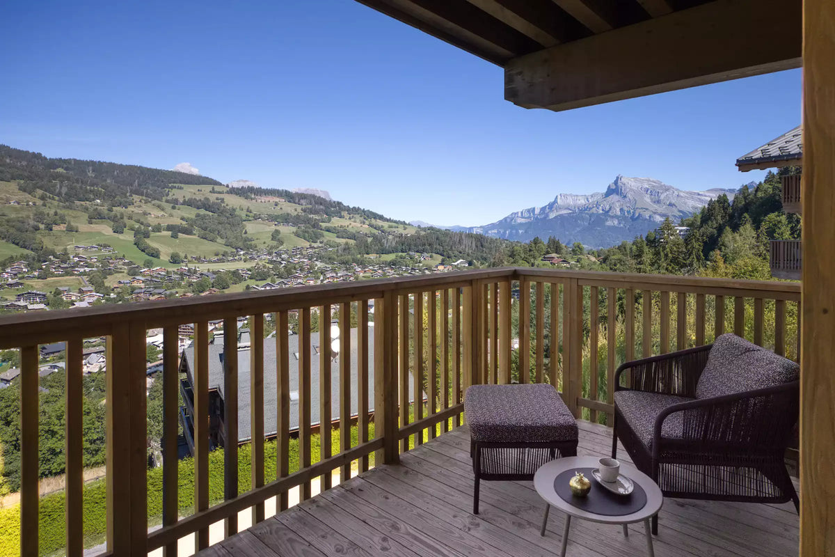 A wooden balcony with a wicker armchair, ottoman, and small round table, overlooking a scenic mountain valley and green hills under a clear blue sky.