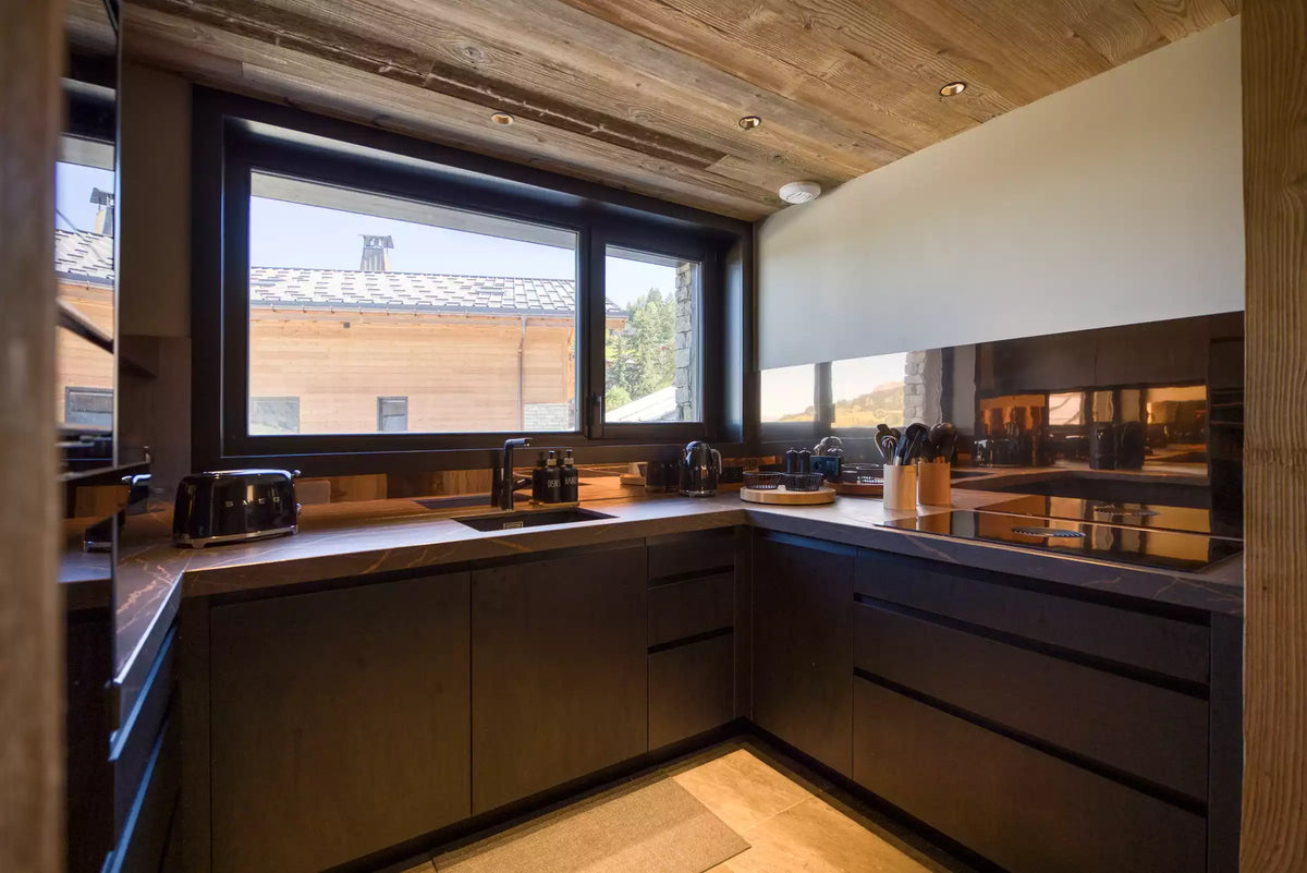 A modern L-shaped kitchen with dark wood cabinets, a dark wood ceiling, and a large window looking out onto an outdoor patio. The countertops are a warm stone, and the backsplash is a glossy dark wood mirror.