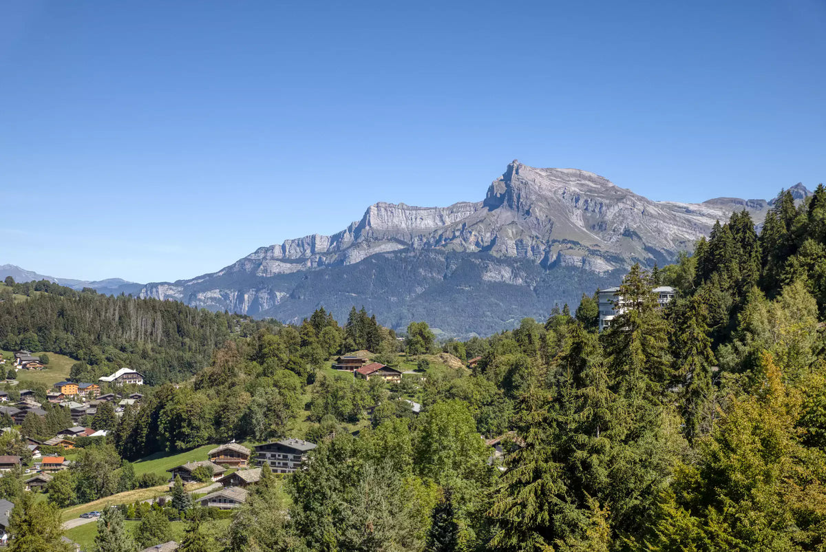 A scenic mountain landscape with a small alpine village nestled in green forested hills, with a large rocky peak rising in the background under a clear blue sky.