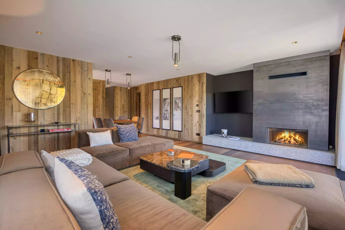 A modern living room featuring a large beige sectional sofa, a low marble-top coffee table, a green patterned rug, and a concrete fireplace wall with a lit fireplace.