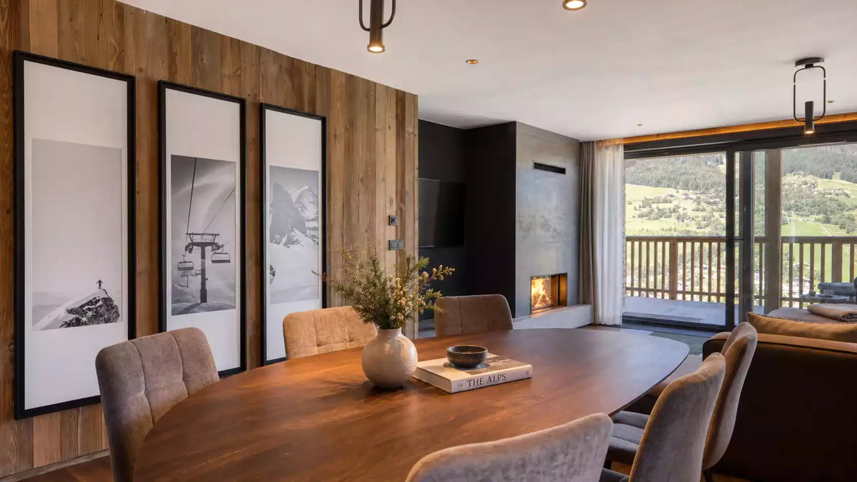 A modern dining room interior with a large wooden dining table and upholstered chairs, set against a wood-paneled wall with three large framed black-and-white artworks. A fireplace, sliding glass doors, and a balcony overlooking a mountain landscape are visible.