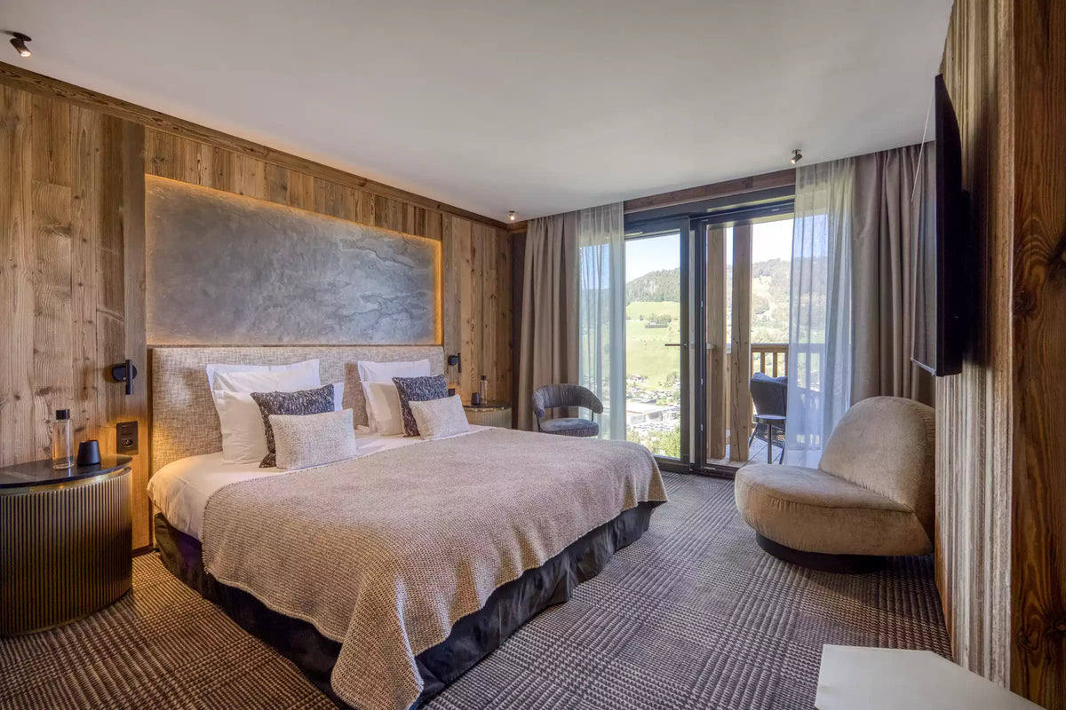 A luxury mountain-style bedroom featuring a large bed with beige bedding, a textured wood headboard and walls, a modern curved armchair, and floor-to-ceiling glass doors overlooking a scenic outdoor view.