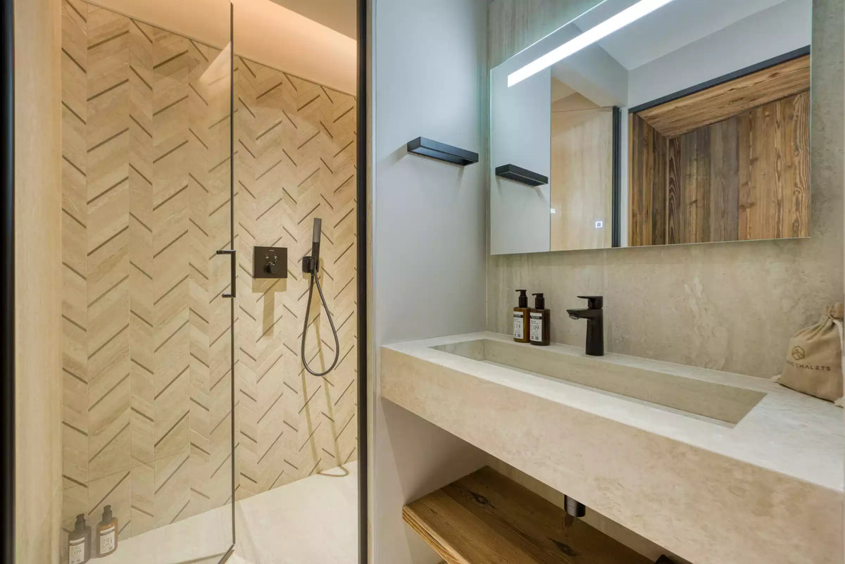 Modern bathroom interior featuring a glass-enclosed shower with beige herringbone tile walls, a floating stone sink vanity, black faucet and hardware, and a large illuminated mirror.