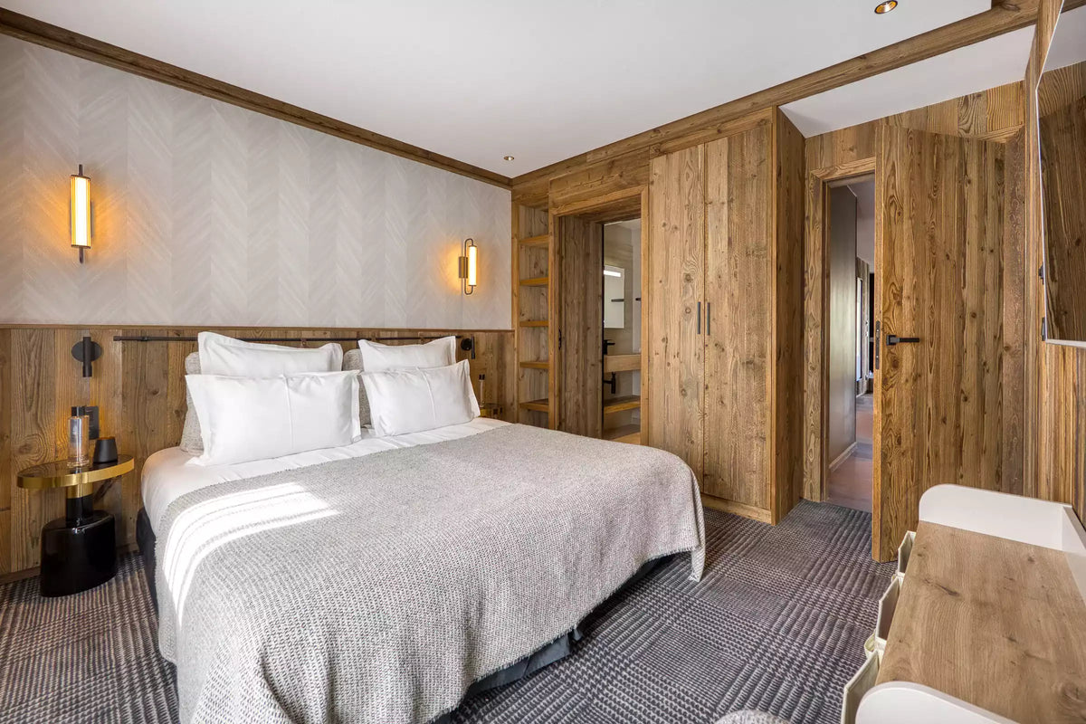 A modern bedroom interior featuring a neatly made bed with white linens and a light gray textured cover, surrounded by warm wood paneling, wall sconces, built-in shelving, and an open doorway revealing more of the room.