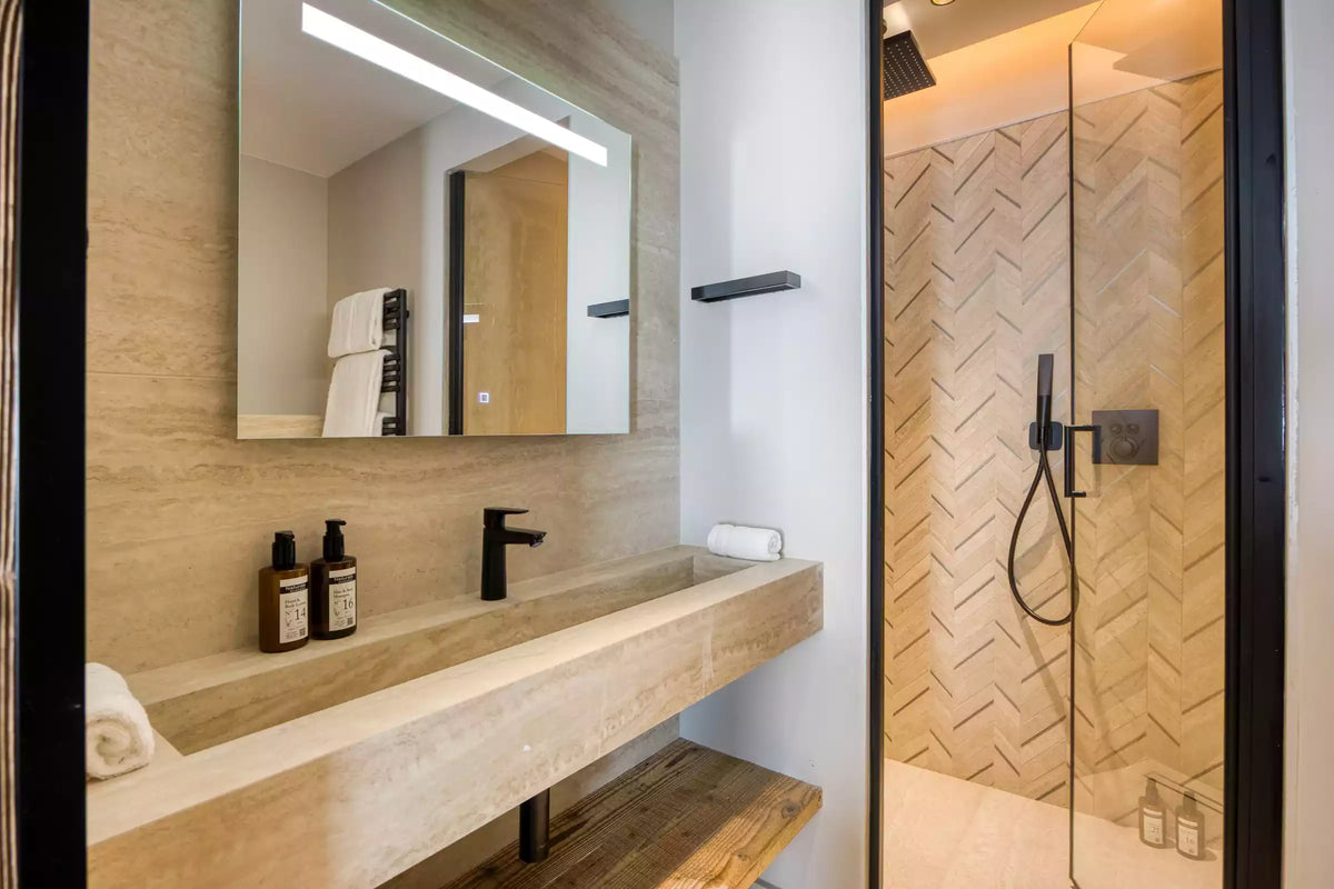 Modern bathroom vanity area with a large rectangular mirror, built-in stone sink, black faucet, and a glass-enclosed shower stall with beige herringbone tile.