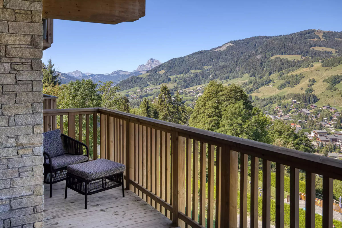A wooden balcony with a stone pillar and two dark outdoor armchairs with cushions, overlooking a lush green mountain valley and village under a clear blue sky.