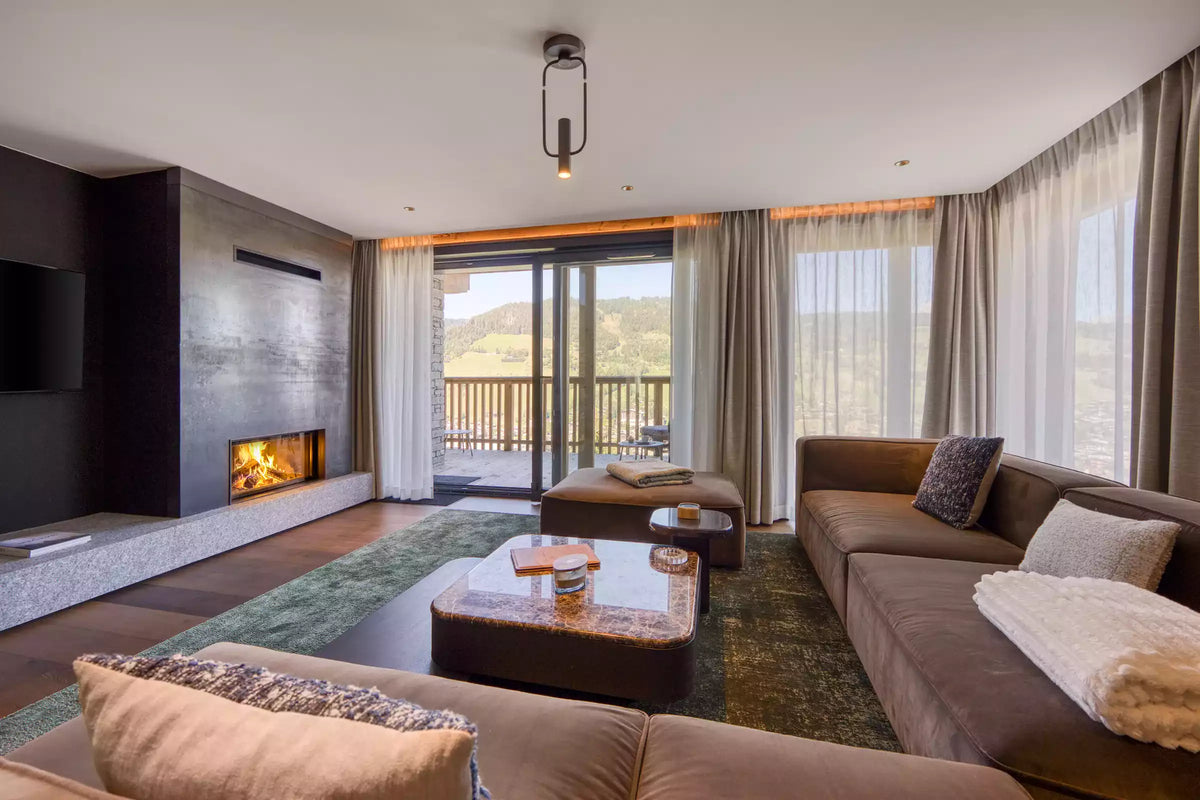 A modern living room with brown sectional sofas, a low rectangular coffee table, a green patterned area rug, a dark wood fireplace with a lit fire, and large windows looking out onto a mountain view.
