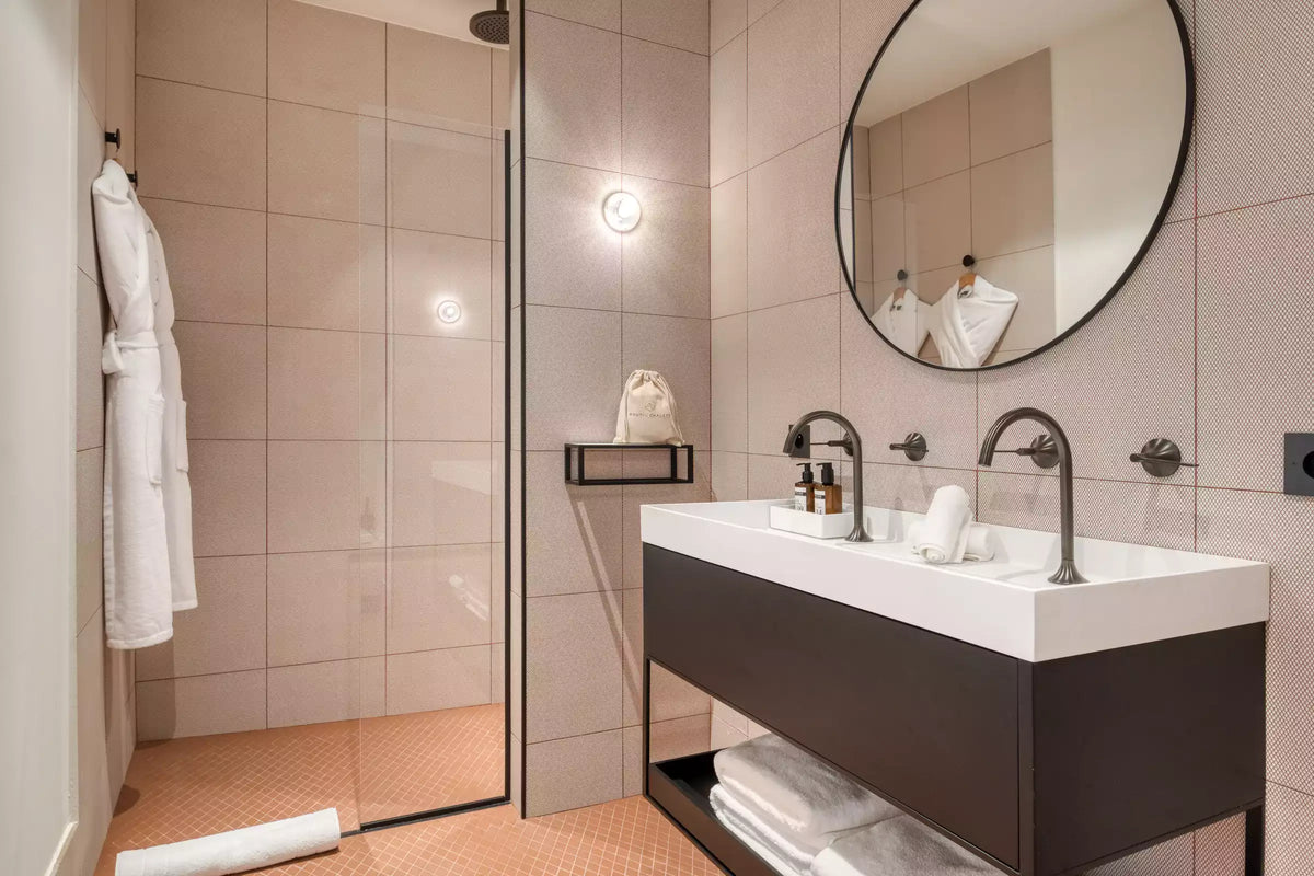 Modern bathroom vanity with a white rectangular sink, dark floating cabinet, round wall mirror, and walk-in glass shower enclosure with beige tiled walls.