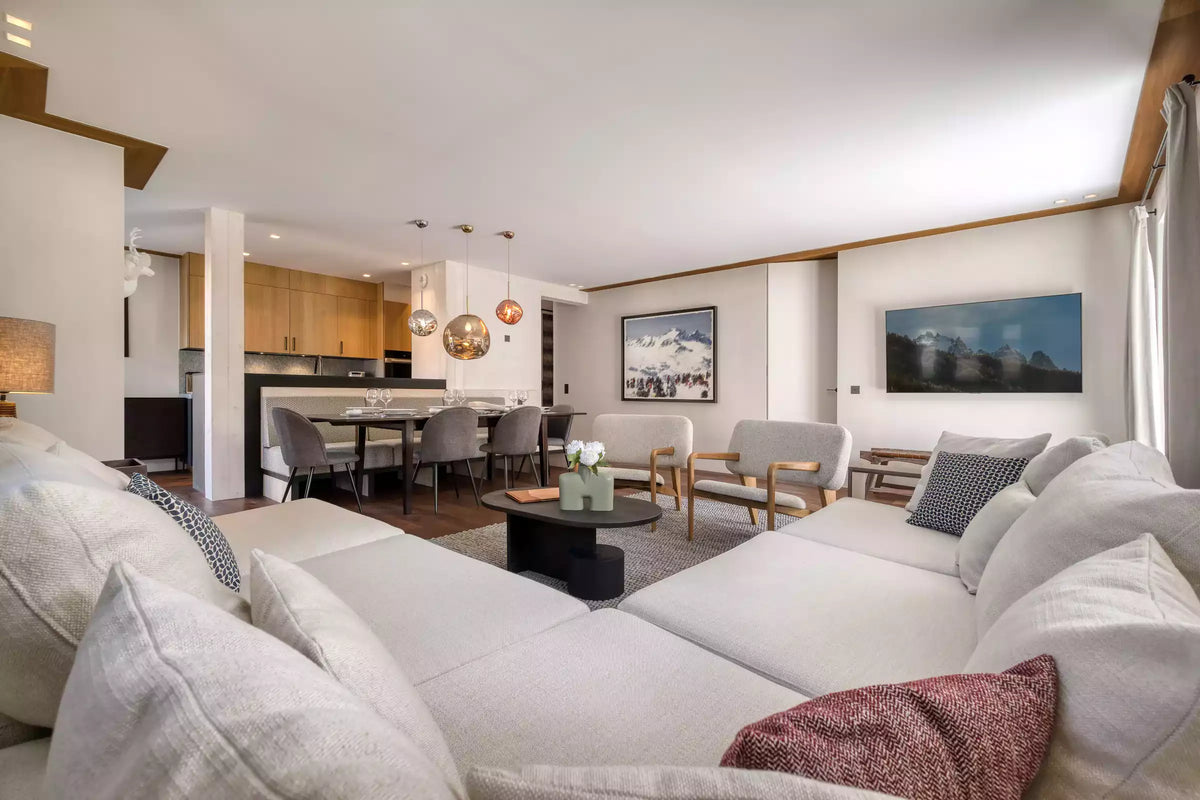 A modern open-plan living room featuring a large white sectional sofa in the foreground, a black round coffee table, light gray armchairs, and a dining area in the background.